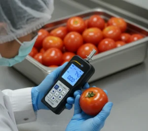 Operator QC Indonesia sedang mengukur kekokohan tomat menggunakan PCE-PTR 200N dengan penetration tip Ø8 mm di area packing house; angka gaya puncak tampil di layar LCD, sampel lain tersusun di baki stainless.