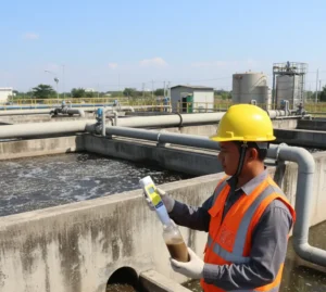 Teknisi Indonesia memegang ECscan10H Pocket Conductivity Tester di tepi bak pengolahan air limbah industri, sensor alat dicelupkan ke dalam botol sampel untuk mengukur konduktivitas air proses secara cepat dan akurat.