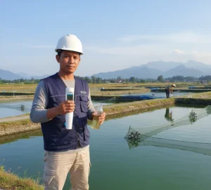 Teknisi Indonesia sedang mengukur pH air kolam menggunakan S10 Bluetooth pH Tester di lapangan, dengan tampilan angka pH yang jelas pada layar alat dan latar belakang tambak ikan.