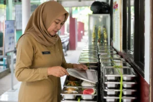 Guru memeriksa makanan dalam wadah stainless steel pada Program Makan Bergizi Gratis (MBG) sebagai bagian dari pengawasan keamanan pangan sebelum didistribusikan.