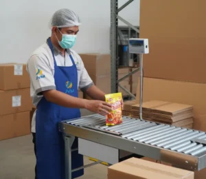 Operator QC Indonesia memeriksa berat kemasan snack di atas checkweighing scale roller PCE-SD 30CR, melihat display LCD berkabel di area packing UMKM.
